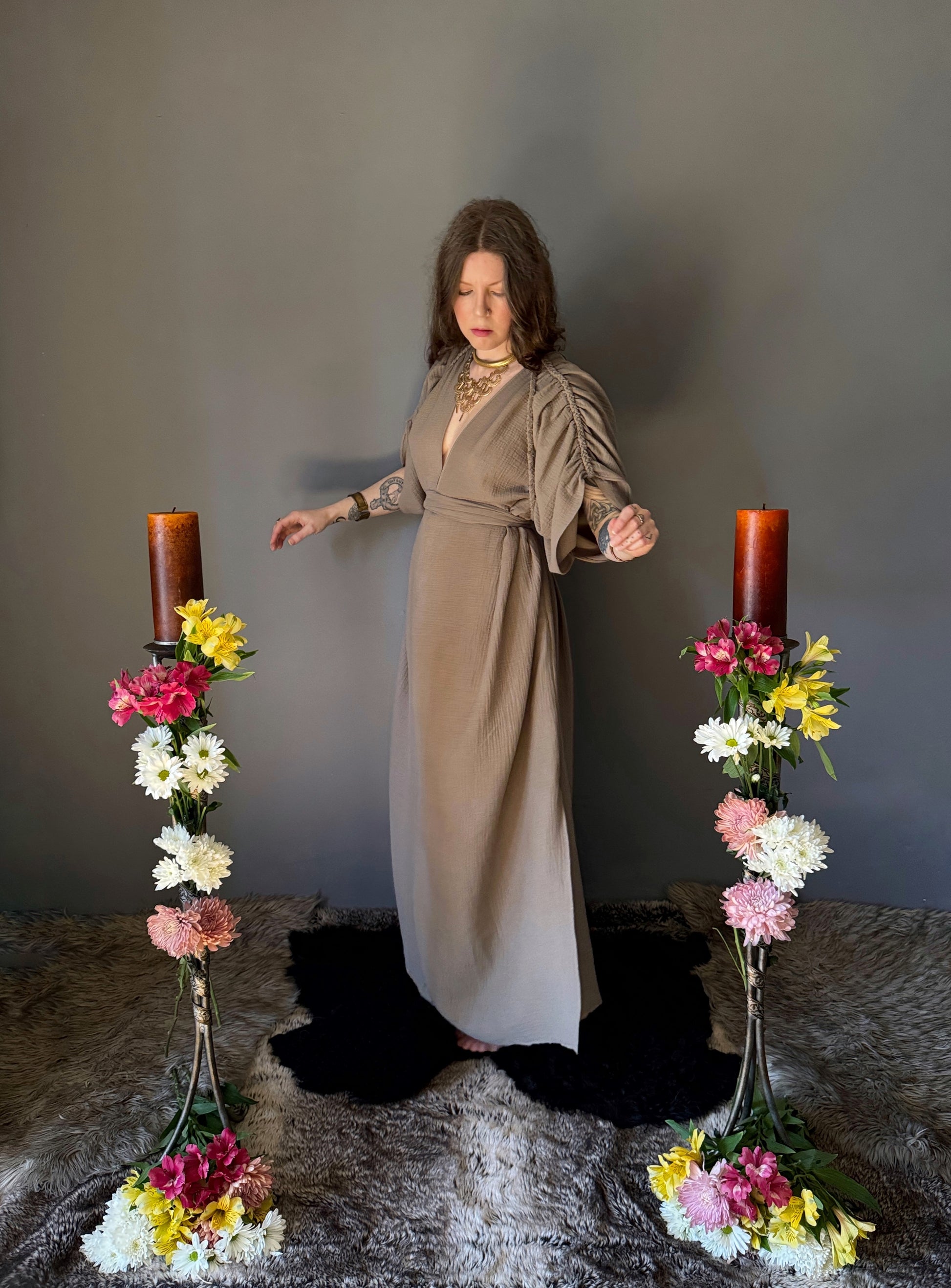 A priestess surrounded by flowers wearing a ancient greek inspired ritual robe. Featuring a deep v neck line, gathered short sleeves with braided trim that ties at the back. Cinched at the waist with a matching long wrap belt. The gown is a soft tan colour cotton with a slight waffle comb texture. The veil is made from a cotton gauze in a natural undyed state. For the magically inclined, witches, priestess, priest, pagan, hellenistic, polytheistic, spiritual or lover of history. Sustainably made in Canada