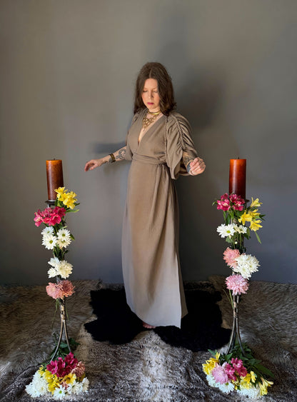 A priestess surrounded by flowers wearing a ancient greek inspired ritual robe. Featuring a deep v neck line, gathered short sleeves with braided trim that ties at the back. Cinched at the waist with a matching long wrap belt. The gown is a soft tan colour cotton with a slight waffle comb texture. The veil is made from a cotton gauze in a natural undyed state. For the magically inclined, witches, priestess, priest, pagan, hellenistic, polytheistic, spiritual or lover of history. Sustainably made in Canada