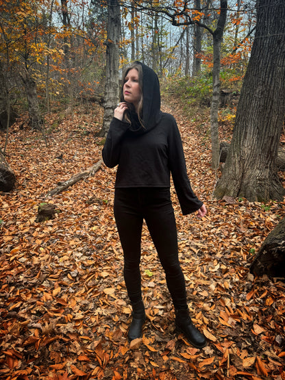 A witch standing in a fprest covered in fallen leaves wearing black pants and a black hexen hoodie. The hoodie features a large cowl hood and cropped just at top of the hips. Featuring slight bell sleeves and its made from a soft bamboo knit with a fleece interior. Very soft and warm good for layering. Witch clothing, fall wear, sustainably made in canada
