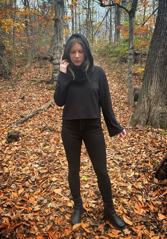 A witch standing in a fprest covered in fallen leaves wearing black pants and a black hexen hoodie. The hoodie features a large cowl hood and cropped just at top of the hips. Featuring slight bell sleeves and its made from a soft bamboo knit with a fleece interior. Very soft and warm good for layering. Witch clothing, fall wear, sustainably made in canada