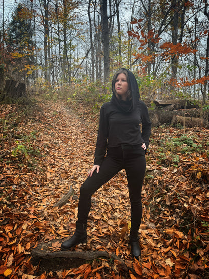 A witch standing in a fprest covered in fallen leaves wearing black pants and a black hexen hoodie. The hoodie features a large cowl hood and cropped just at top of the hips. Featuring slight bell sleeves and its made from a soft bamboo knit with a fleece interior. Very soft and warm good for layering. Witch clothing, fall wear, sustainably made in canada