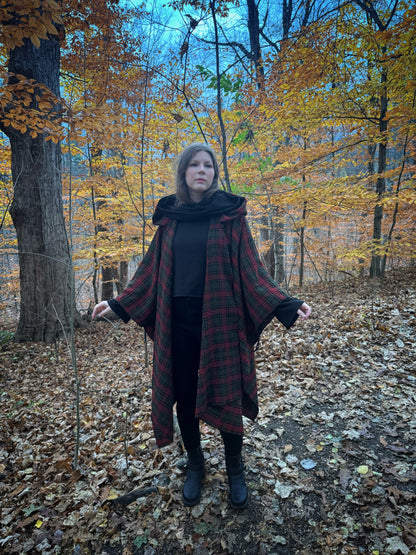 A witch standing in a forest covered in fallen leaves wearing black pants and a black hexen hoodie with a tartan poncho layered over top, the hood is down featuring the cowl. The hoodie features a large cowl hood and cropped just at top of the hips. Featuring slight bell sleeves and its made from a soft bamboo knit with a fleece interior. Very soft and warm good for layering. Witch clothing, fall wear, sustainably made in canada