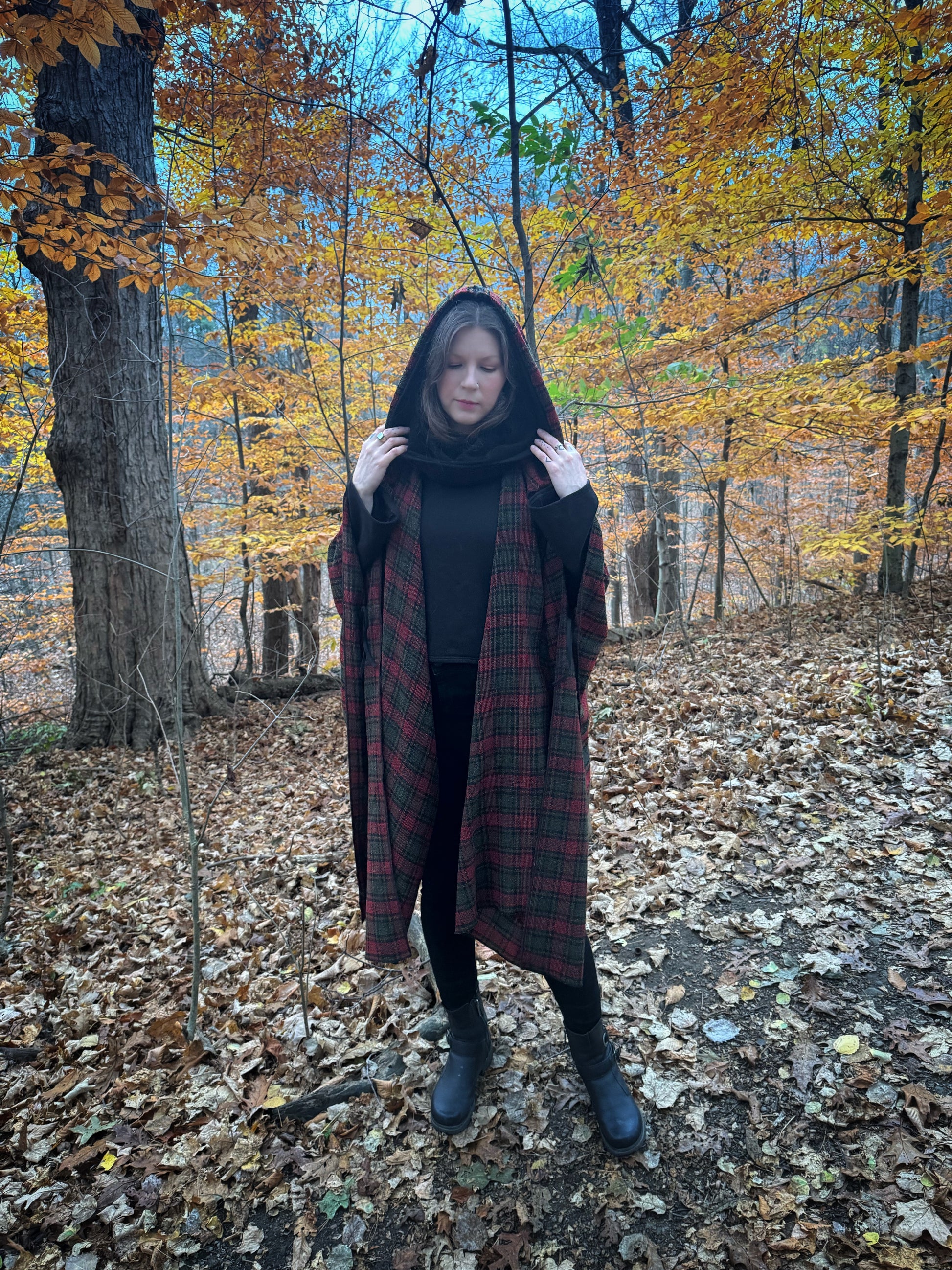 A witch standing in a fprest covered in fallen leaves wearing black pants and a black hexen hoodie with a tartan poncho layered over top. The hoodie features a large cowl hood and cropped just at top of the hips. Featuring slight bell sleeves and its made from a soft bamboo knit with a fleece interior. Very soft and warm good for layering. Witch clothing, fall wear, sustainably made in canada