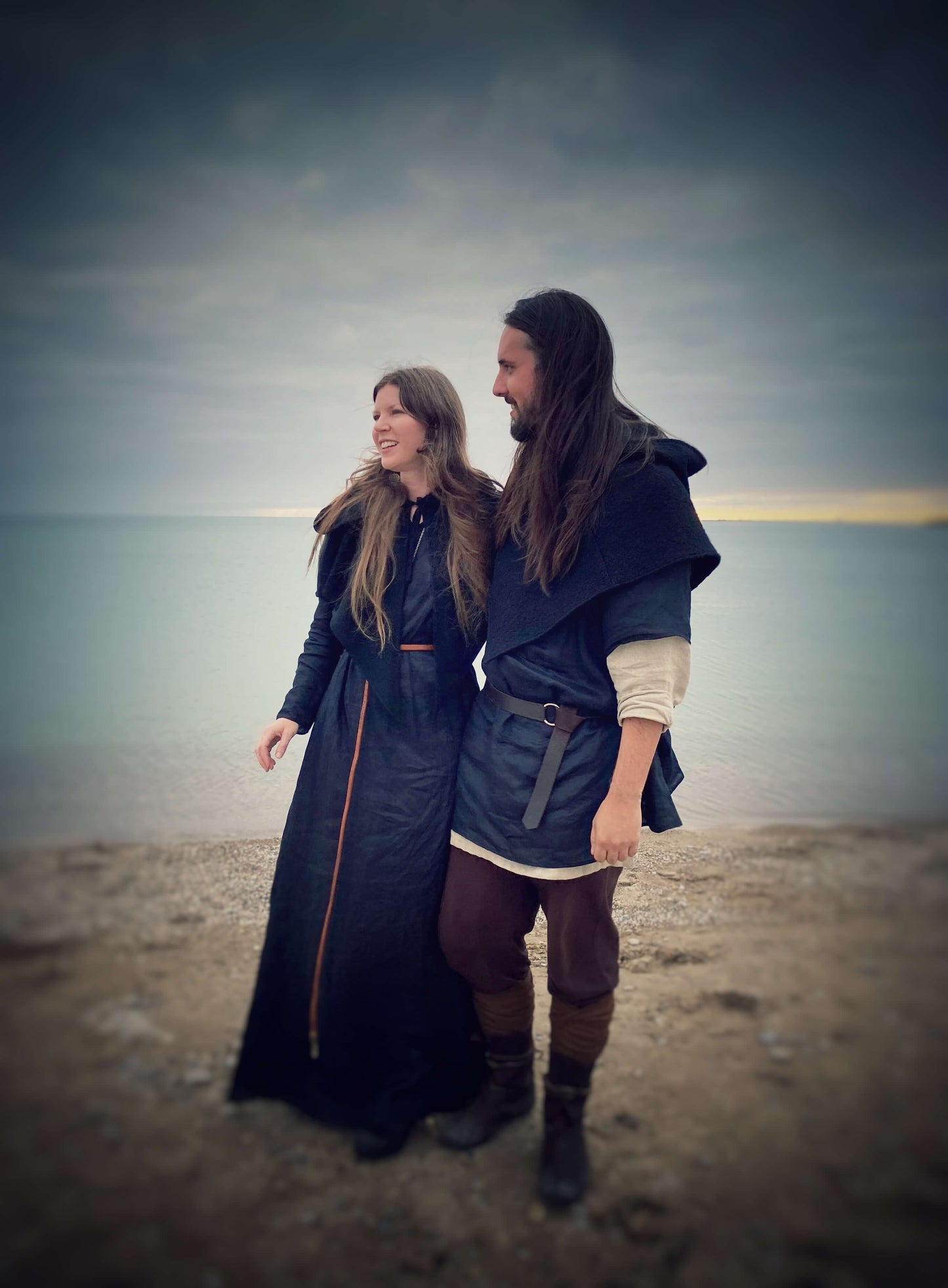 Female model wearing the Bridei's tunic dress in black linen paired witha wool capelet open at the front. The male model is wearing a norse costume with one of our blac wool norse hoods.