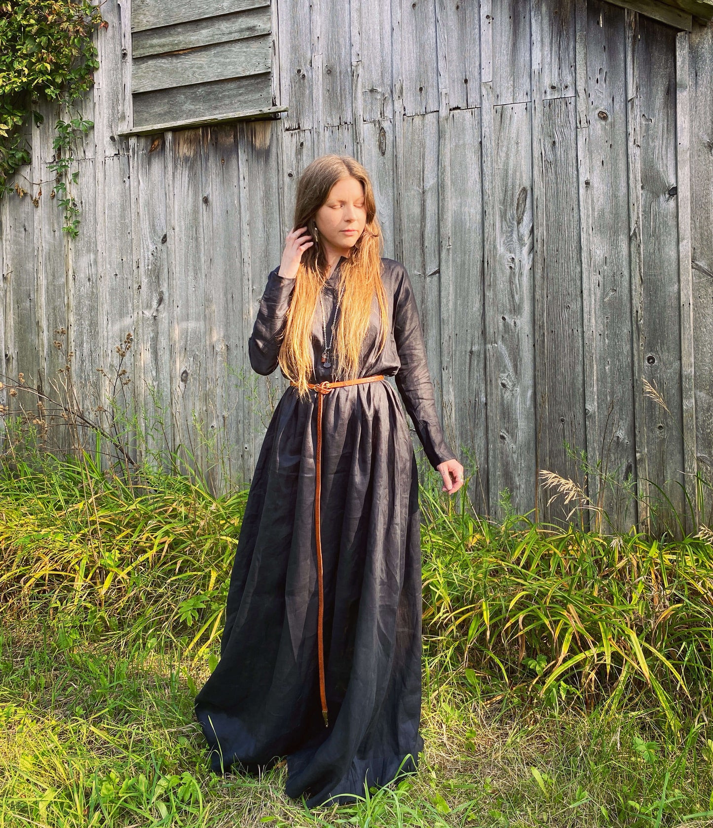 Featuring our Iron age, long sleeve tunic dress in black linen. Model is wearing it with a thin leather belt with a barn in the background.