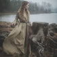 This photo features our iron age tunic dress in a tan colour. The model is wearing a leather belt, holding a sword with a lake in the background.