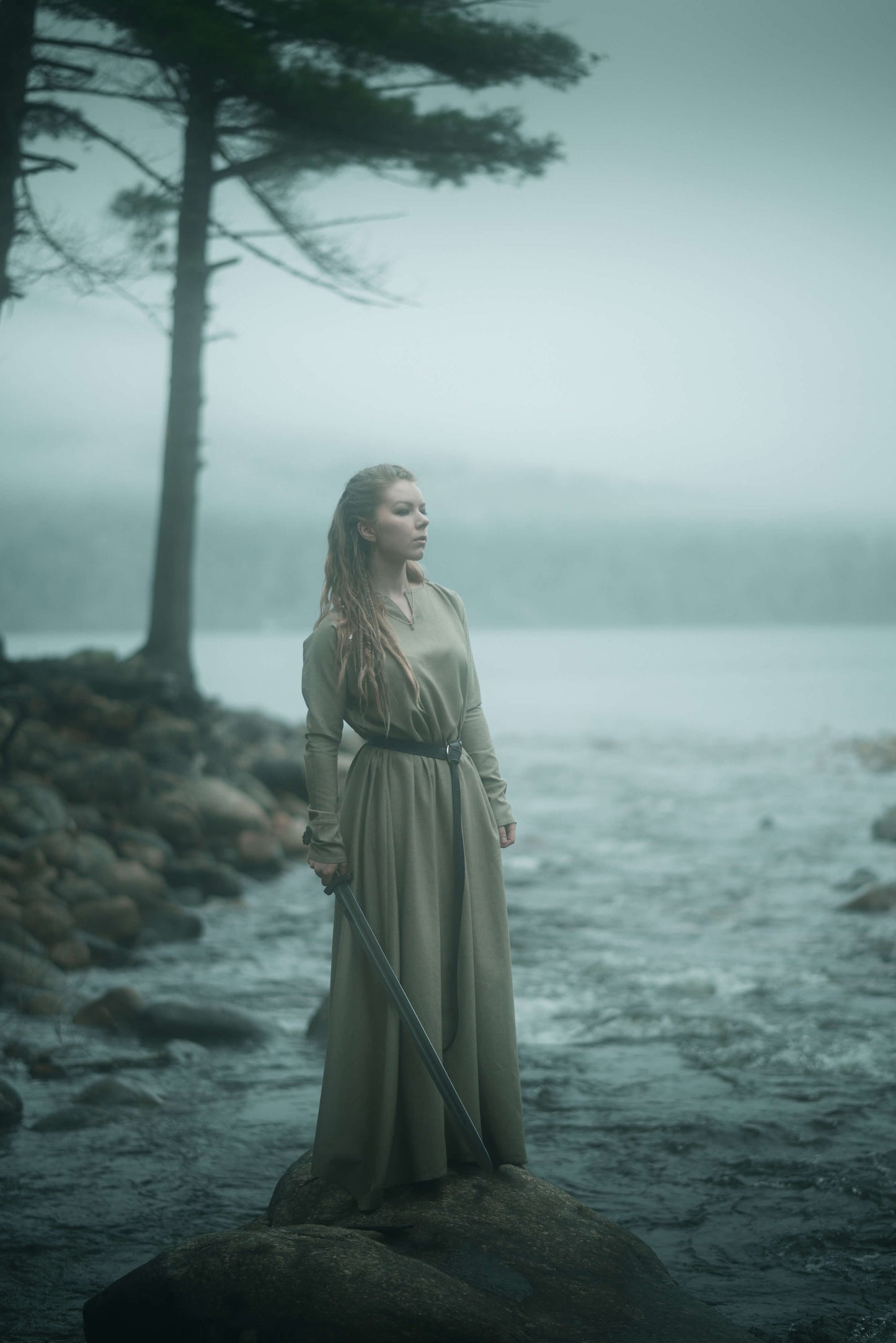 Model is wearing our long sleeve, tunic dress in a natural colour. Standing on a rock, holding a sword with a lake in the background.