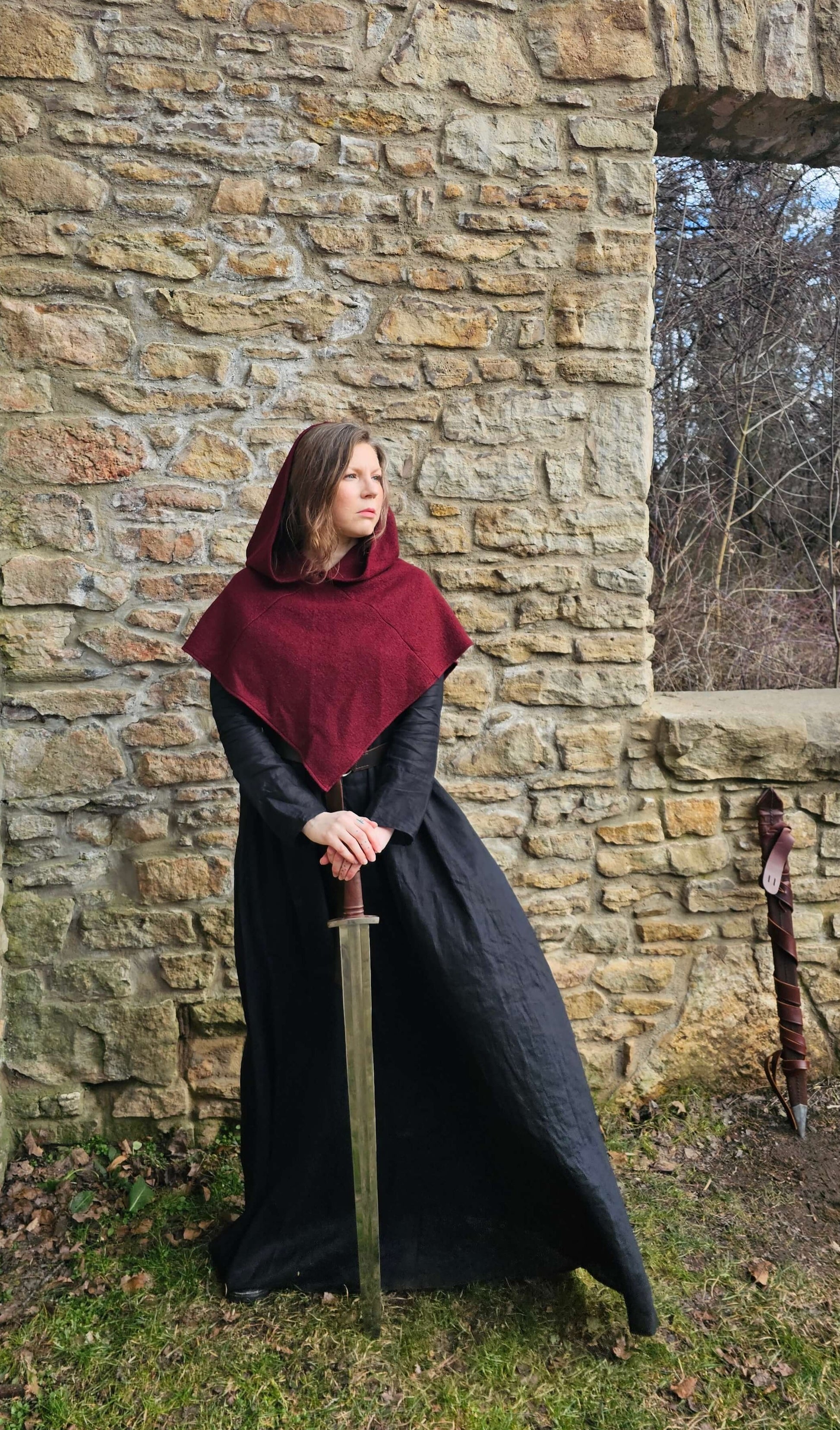 A woman standing, looking to the side while holding a sword she has propped in front of her. The wind is blowing her viking inspired, Bridei's Tunic dress  paired with a burgundy woold Norse Hood.  She is standing proudly infront of a stone wall. 