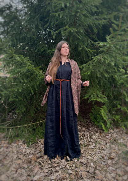 A celtic woman stands in front  some evergreen trees wearing a black tunic dress with a brown long belt and featuring an earth toned plaid shawl. Brown undertones with white and red stripes. Just draped over the shoulders, open front in this photo.
Historical costume, ancestor reverence, old gods, made in canada.