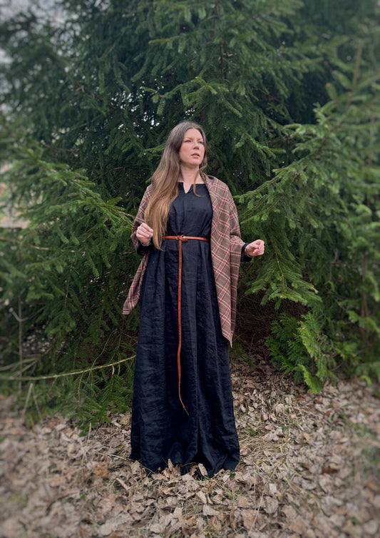 A celtic woman stands in front  some evergreen trees wearing a black tunic dress with a brown long belt and featuring an earth toned plaid shawl. Brown undertones with white and red stripes. Just draped over the shoulders, open front in this photo.
Historical costume, ancestor reverence, old gods, made in canada.