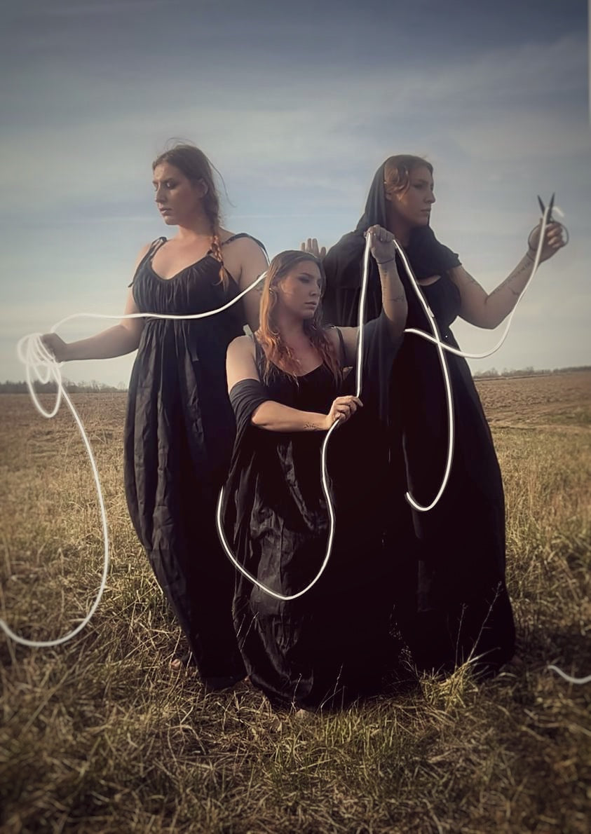 In an open feild stand the three fates all wearing the black gown and black priestess veil weaving the string of fate. The fates gown features adjuastble straps that tie at the shoulder, side seam pockets and a long wrap belt to tie at the waist. Priestess, ritual gown, sustainable clothing, made in canada